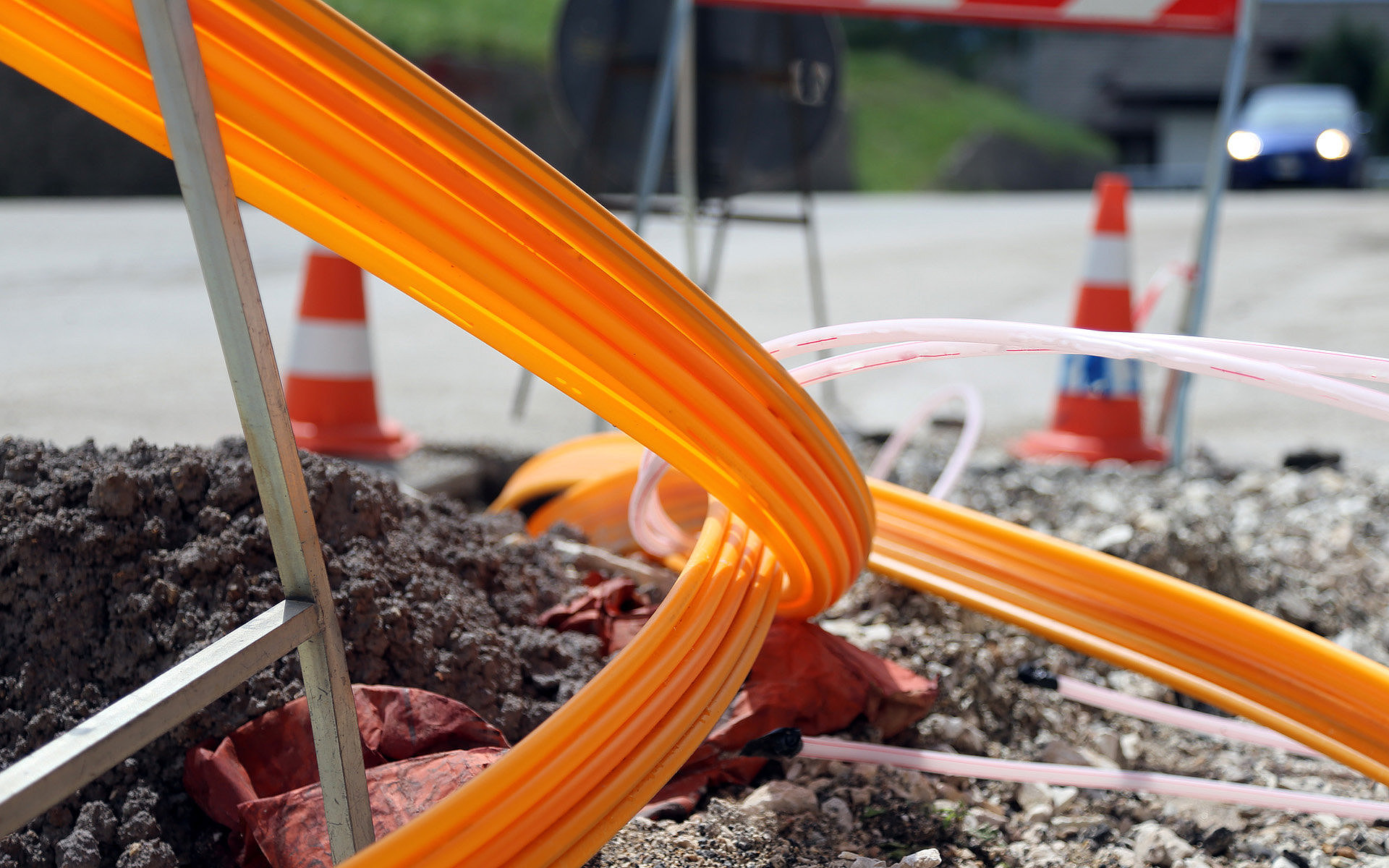 Straßenbaustelle mit leuchtend orangefarbenen Leerrohren, die aus einem aufgegrabenen Schacht hervorragen, umgeben von Schotter und Warnkegeln.